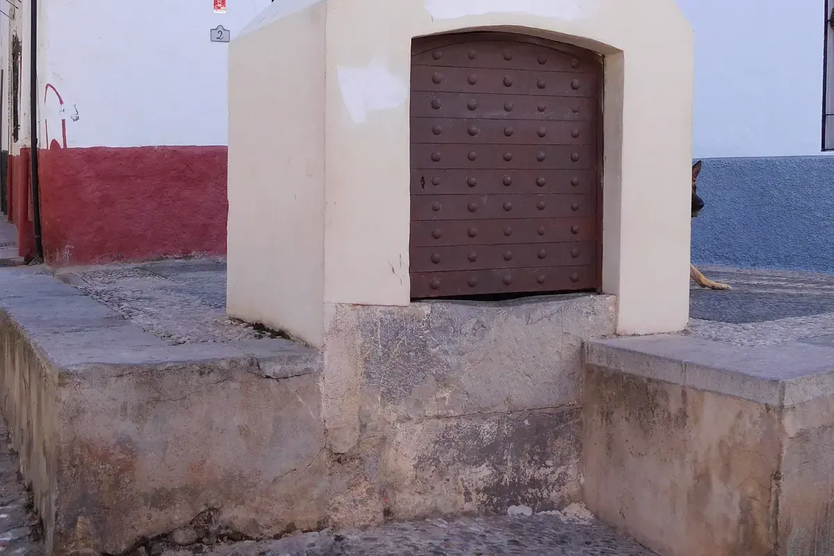 A whitewashed stone cistern with a studded wooden door on Calle Panaderos in the Albaicín, beside a red-painted wall and a stone step