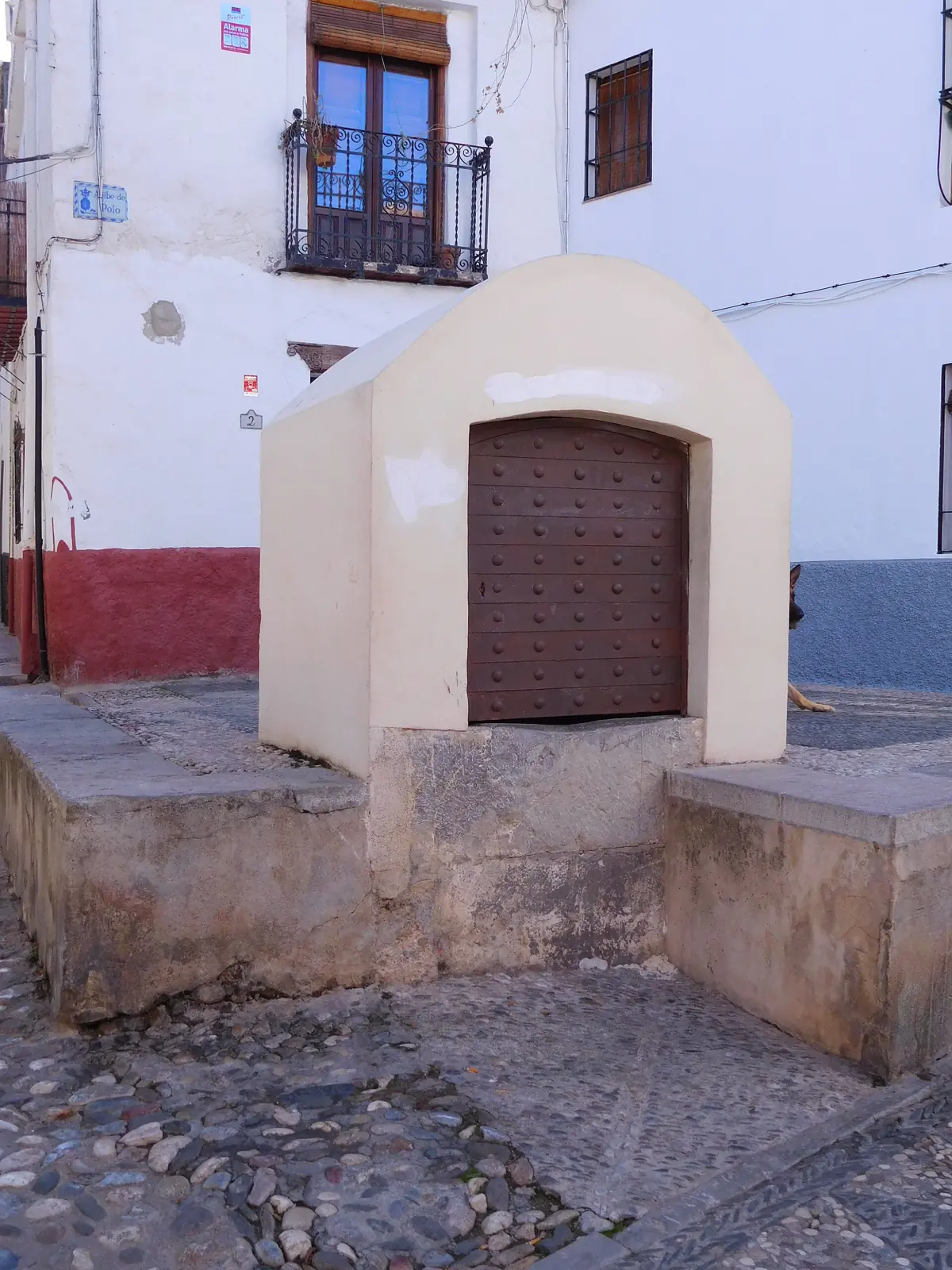 A whitewashed stone cistern with a wooden door on Calle Panaderos in the Albaicín, beside a balcony and a cobbled corner